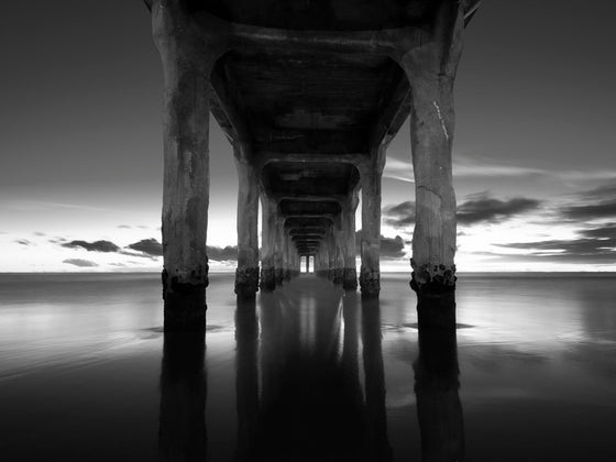 Manhattan beach pier at sunset with blue skies and a reflection of the pier and clouds in the sand at low tide, in black and white