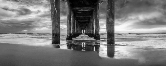 A panoramic photograph of underneath the Manhattan Beach pier during sunset, in black and white