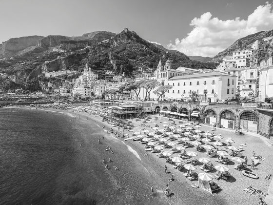 Black and white image of the beautiful Amalfi town on the Amalfi coast in Italy with water, beach clubs and umbrellas with the mountains in the background.