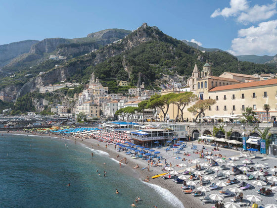 Amalfi Beach Town with beach umbrellas at the Amalfi Beach Club with the Mediterranean ocean in the front left corner and some of the mountain hills in the background.