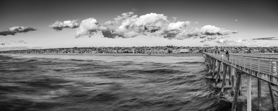 A photo of the city of Hermosa Beach and the Hermosa Beach pier taken at the end of the pier. Pink clouds are illuminated over the houses and the water in black and white.