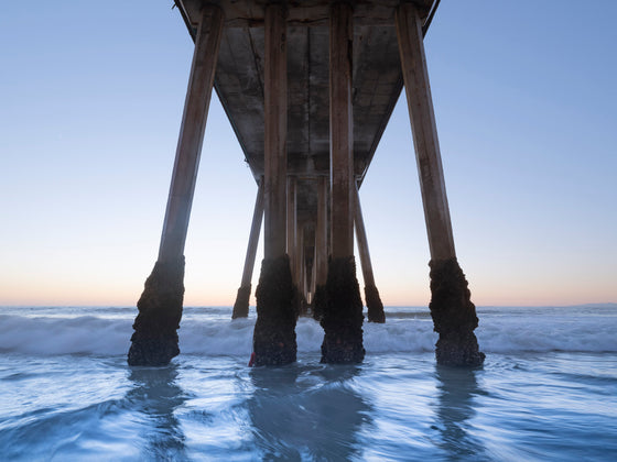 Hermosa Beach pier during golden hour. The barnacles on the pillars of the pier are the focal point as the light blue water sways against them and the orange horizon is seen behind them.