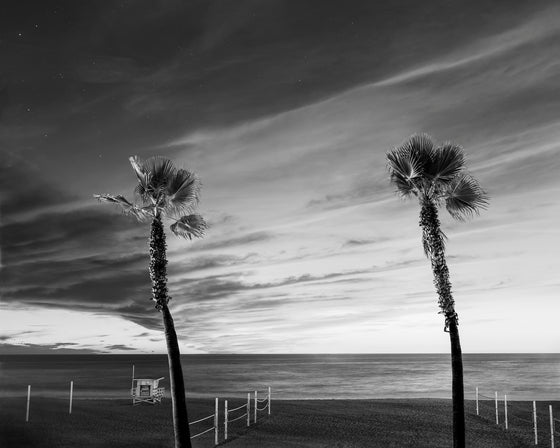manhattan beach sunset, with clouds and a sky with stars, two palm trees, volleyball courts and a lifeguard tower, in black and white
