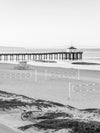 Sunrise over the Pacific Ocean at the Manhattan Beach Pier, with surfers and a small swell and decent surf, with a beach cruiser, volleyball courts and a lifeguard tower in the foreground, black and white