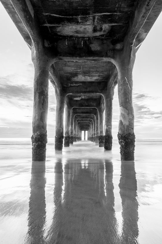 A vertical black and white photograph from under the Manhattan Beach pier during low tide