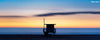 A panoramic photo of a lifeguard tower in Hermosa Beach / Manhattan Beach (Los Angeles California) at sunset, with blue and yellow skies, as golden hour fades into the blue hour at the beach over the ocean.