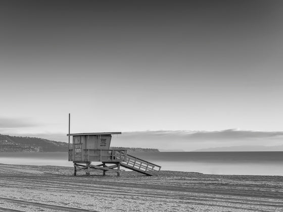 A horizontal picture of a lifeguard tower in Redondo Beach with Palos Verdes in the background, in black and white