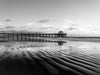 Black and white photo of the Manhattan Beach pier at a very low tide, with repeating ripples of sand in the foreground.