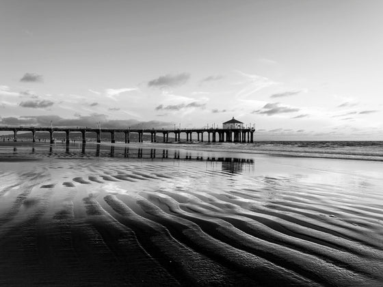Black and white photo of the Manhattan Beach pier at a very low tide, with repeating ripples of sand in the foreground.