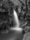 Photo of a waterfall in the Julian Alps, Slovenia. This is a long exposure of a waterfall flowing into a blue pool of water surrounded by rocks on all sides, making the scene appear almost like water flowing into a calm river in black and white.