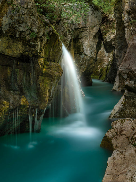 Photo of a waterfall in the Julian Alps, Slovenia. This is a long exposure of a waterfall flowing into a blue pool of water surrounded by rocks on all sides, making the scene appear almost like water flowing into a calm river.