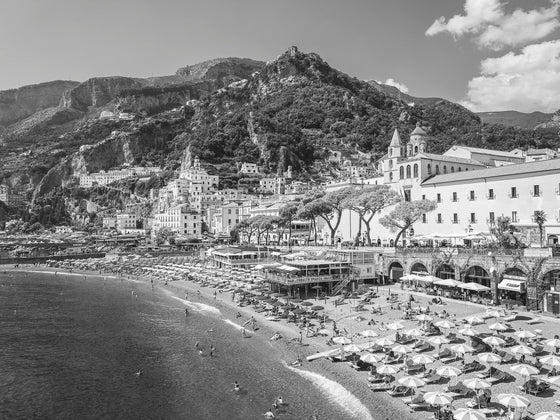 Amalfi Beach Town with  black and white beach umbrellas at the Amalfi Beach Club with the Mediterranean ocean in the front left corner and some of the mountain hills in the background.