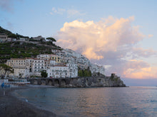  Amalfi Coast houses on the hillside with big pink and white clouds with the Mediterranean Ocean in the foreground.