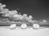A photo of a an empty Caribbean beach with crystal-clear water with three white umbrellas and twelve day beds. The sky is clear with puffy white clouds in it .It's a black and white image.