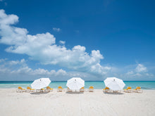  A photo of a an empty Caribbean beach with crystal-clear blue water with three white umbrellas and twelve yellow day beds. The sky is blue and there are puffy white clouds in it.