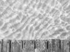 A close-up photo of a dock in the Caribbean with crystal clear water and ripples in the sand below it in black and white.
