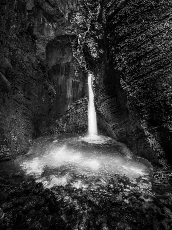 Photo of a waterfall in the Julian Alps, Slovenia. This shows a waterfall flowing into a pool of water surrounded by rocks, making the scene appear almost like water flowing into a cave in black and white.