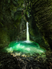  Photo of a waterfall in the Julian Alps, Slovenia. This shows a waterfall flowing into a green pool of water surrounded by rocks, making the scene appear almost like water flowing into a cave