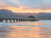 A photograph of Hanalei pier on Kauai during sunset with orange sunrays peaking over a green mountain range. The orange is reflected in the ocean.