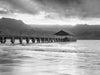 A photograph of Hanalei pier on Kauai during sunset with orange sunrays peaking over a green mountain range. The orange is reflected in the ocean in black and white.