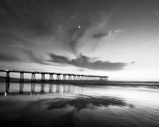 Sunset at Hermosa Beach California pier, low tide, with the clouds reflecting in the sand, and a moon present above the pier, in black and white