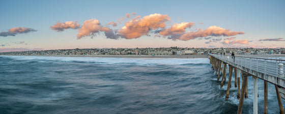 A photo of the city of Hermosa Beach and the Hermosa Beach pier taken at the end of the pier. Pink clouds are illuminated over the houses and the water