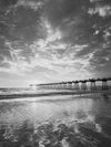 Photo of the Hermosa Beach Pier during sunset with dramatic yellow, orange, and gray clouds and their reflection in the sand in black and white.