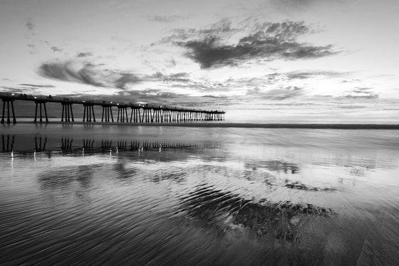 The Hermosa Beach California pier after sunset during the blue hour at low tide with the clouds and sunset reflecting in the ripples in the sand, in black and white
