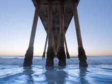 Hermosa Beach pier during golden hour. The barnacles on the pillars of the pier are the focal point as the light blue water sways against them and the orange horizon is seen behind them.