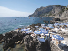  a horizontal picture featuring the blue beach umbrellas of la fontelina beach club in Capri, Italy with the ocean and coastline in the background