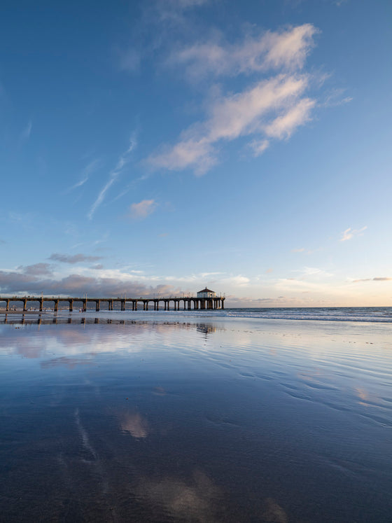 The Manhattan Beach California pier with blue skies and clouds, at low tide, with the clouds reflected in the sand
