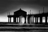 Manhattan Beach Pier during sunset. Dramatic mist and sunrays can be seen through the pillars of the pier. The sky is ombré from purple to pink to orange to yellow. This photo is in black and white.