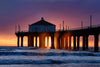 Manhattan Beach Pier during sunset. Dramatic mist and sunrays can be seen through the pillars of the pier. The sky is ombré from purple to pink to orange to yellow.