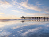 Photo of the Manhattan Beach Pier right before sunset. The clouds are white and are reflected in the sand. The horizon is yellow and the sky and sand are blue. 