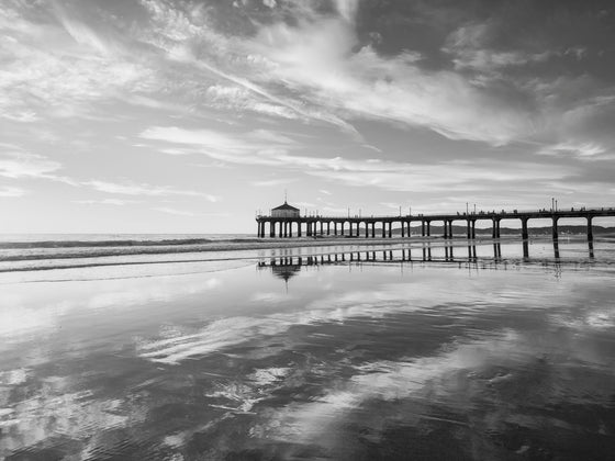 Photo of the Manhattan Beach Pier right before sunset. The clouds are white and are reflected in the sand. The horizon is yellow and the sky and sand are blue in black and white.