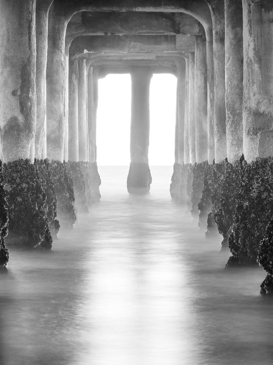 Photo of sunset from underneath the Manhattan Beach pier. It is a long exposure and there is a gold reflection on the water in black and white.