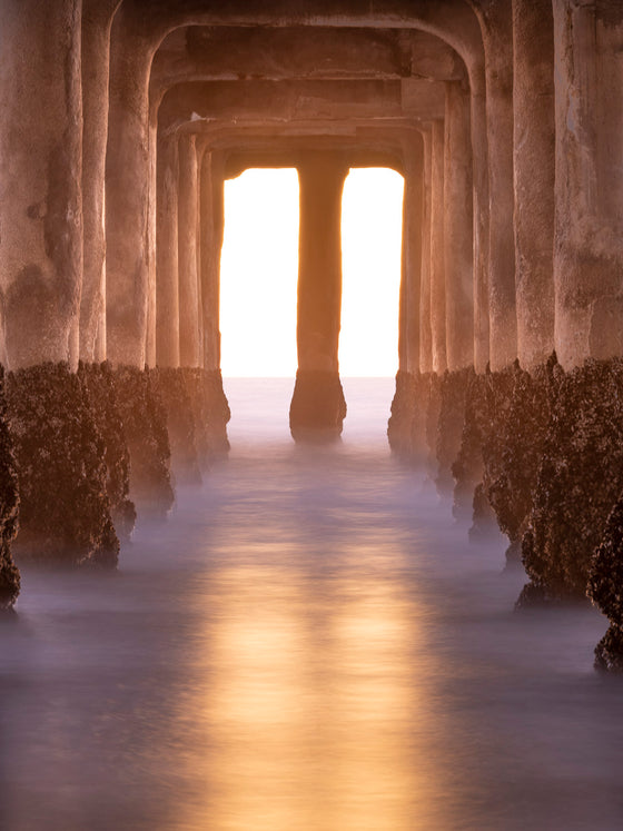 Photo of sunset from underneath the Manhattan Beach pier. It is a long exposure and there is a gold reflection on the water.
