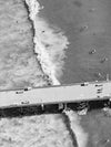 Black and white aerial photo of Manhattan Beach Pier in Los Angeles with surfers, swimmers, and a wave breaking in the Pacific Ocean