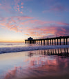 A sunset photo taken on the south side of the Manhattan Beach Pier. The clouds in the sky are pink and orange. They look like a spider with a bunch of legs reaching out for something. The clouds are reflected in the sand because it is low tide.