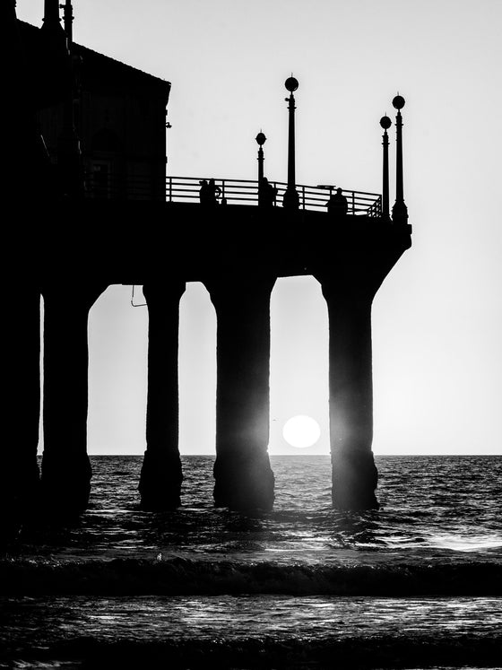 Manhattan Beach sunset shining through the pier's pilings about to reach the horizon in black and white. 