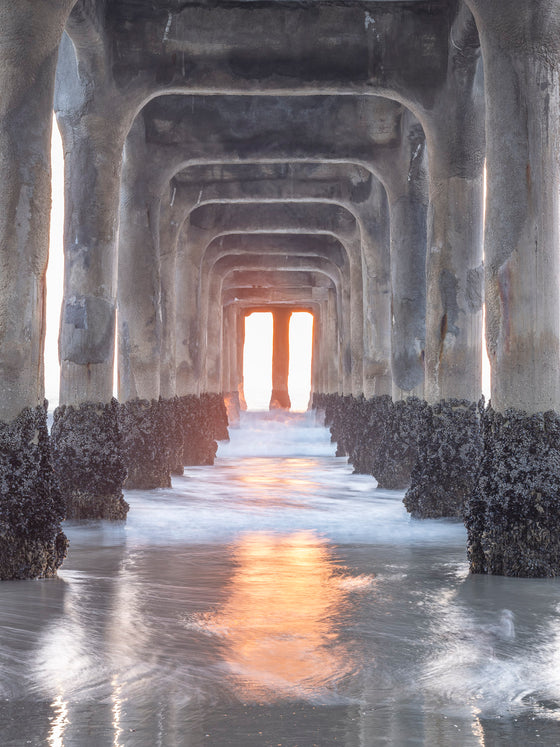 Photo taken under the Manhattan Beach Pier during sunset. There are no clouds in the sky. There is an orange reflection in the sand from the sunset.