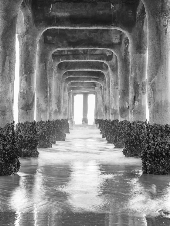 Photo taken under the Manhattan Beach Pier during sunset. There are no clouds in the sky. There is an orange reflection in the sand from the sunset in black and white.