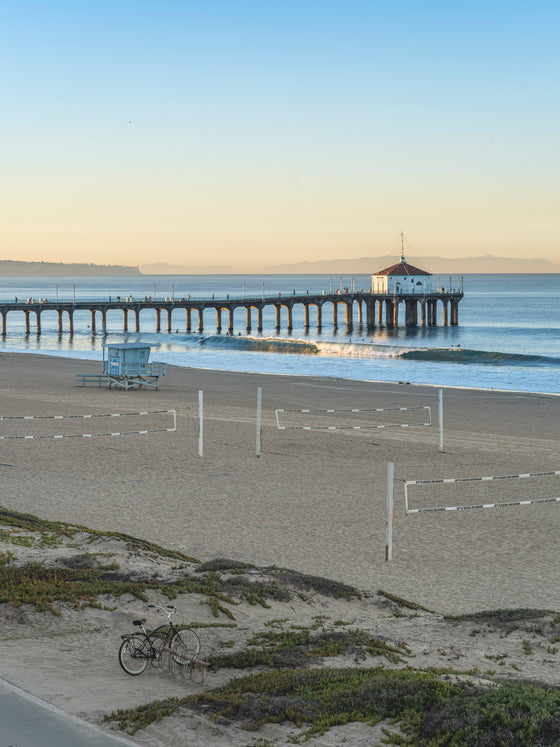 Sunrise over the Pacific Ocean at the Manhattan Beach Pier, with surfers and a small swell and decent surf, with a beach cruiser, volleyball courts and a lifeguard tower in the foreground