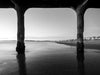 Sunset photo of the Manhattan Beach Pier taken during sunset. Northern Manhattan Beach and the Santa Monica Bay can be seen in this photo. There are blue tones and the sand is reflecting the sky in black and white.
