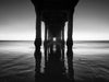 Photo taken under the Manhattan Beach Pier during sunset. There are no clouds in the sky. The sand and the sky are blue. Above the horizon the sky is orange in black and white.