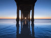 Photo taken under the Manhattan Beach Pier during sunset. There are no clouds in the sky. The sand and the sky are blue. Above the horizon the sky is orange.