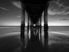 Photo of sunset from underneath the Manhattan Beach pier. It is a long exposure and there are purple clouds while the sky fades from deep blue to orange as it approaches the horizon in black and white.