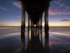 Photo of sunset from underneath the Manhattan Beach pier. It is a long exposure and there are purple clouds while the sky fades from deep blue to orange as it approaches the horizon.