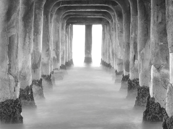 Photo taken from underneath the Manhattan Beach pier. It is a long exposure and the ocean looks flat and there is mist on it in black and white.