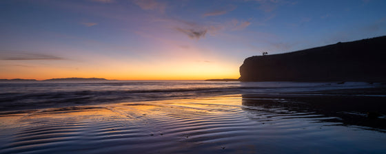 Palos Verdes sunset photo. Catalina Island can be seen on the left above the water and to the right there is a large cliff. The low tide reflects the orange sunset in the sand.